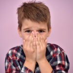 Boy with light brown hair in a plaid shirt covers his mouth because of bad breath in Columbus, OH